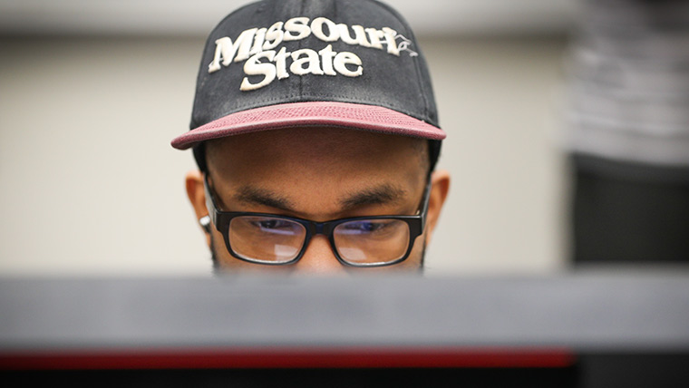 Student looking at computer screen while wearing Missouri State hat.