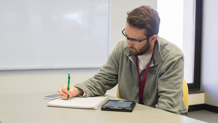 A student writes in a notebook. His tablet computer and phone sit on either side of the notebook.