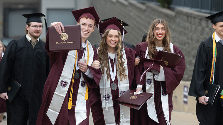 Three students in graduation regalia happily display their diplomas after their commencement ceremony.