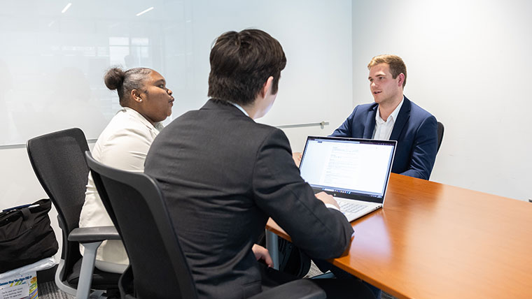 Three professionally-dressed BEAR Essentials students meet for a discussion in a conference room, while one student searches the Internet on a laptop.
