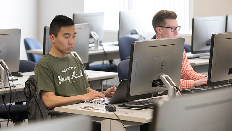 Two students in a computer lab focus on their respective screens as they type.