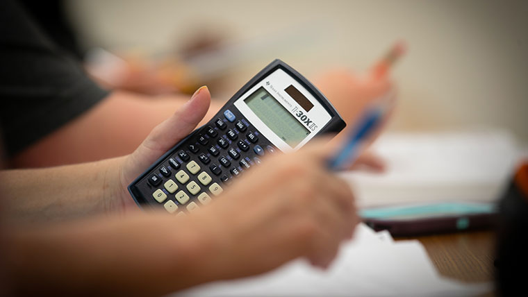 A student's left hand holding a calculator and right hand holding a pencil.